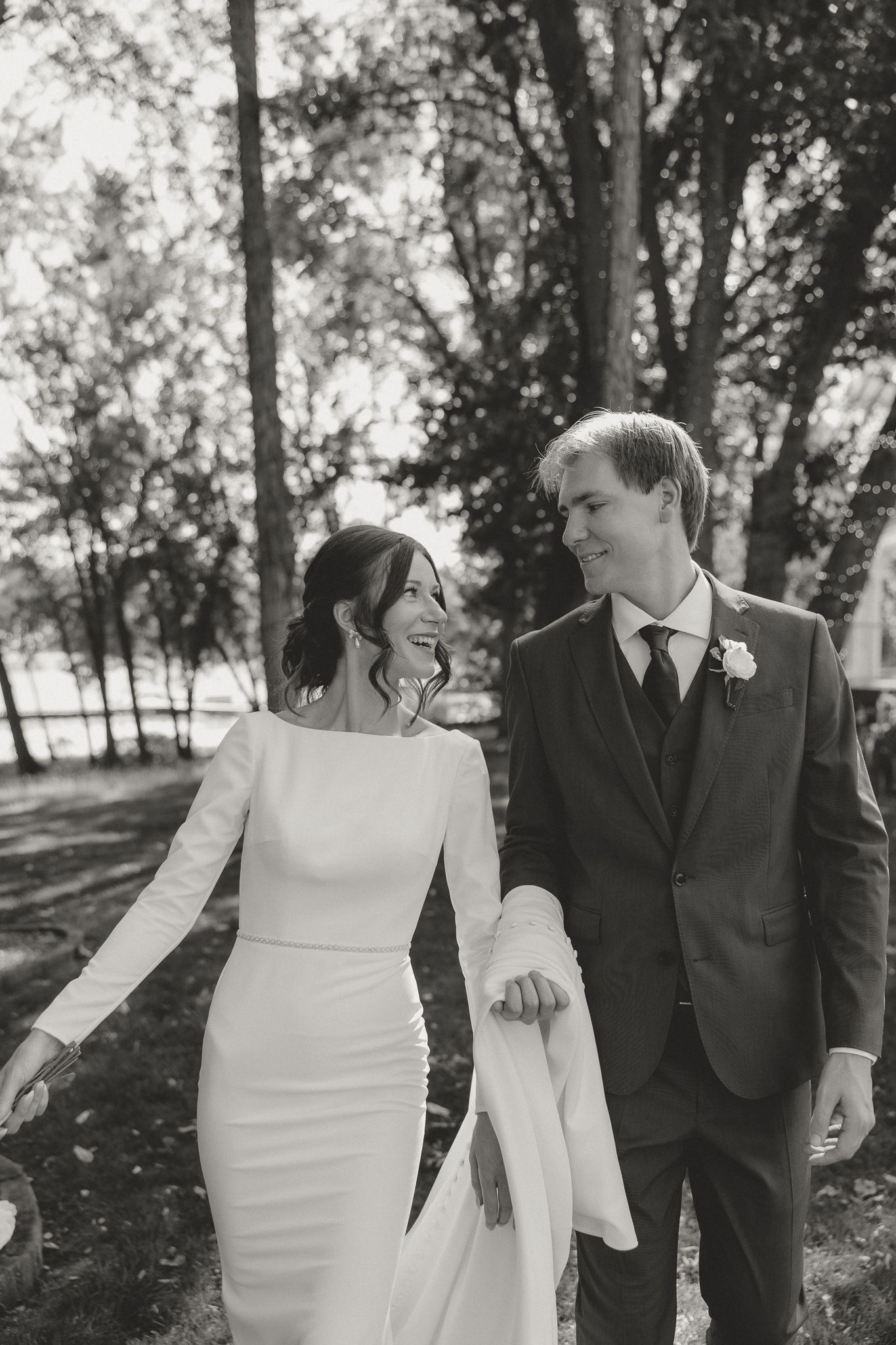 A couple walks together in elegant wedding attire through a tree-lined path.