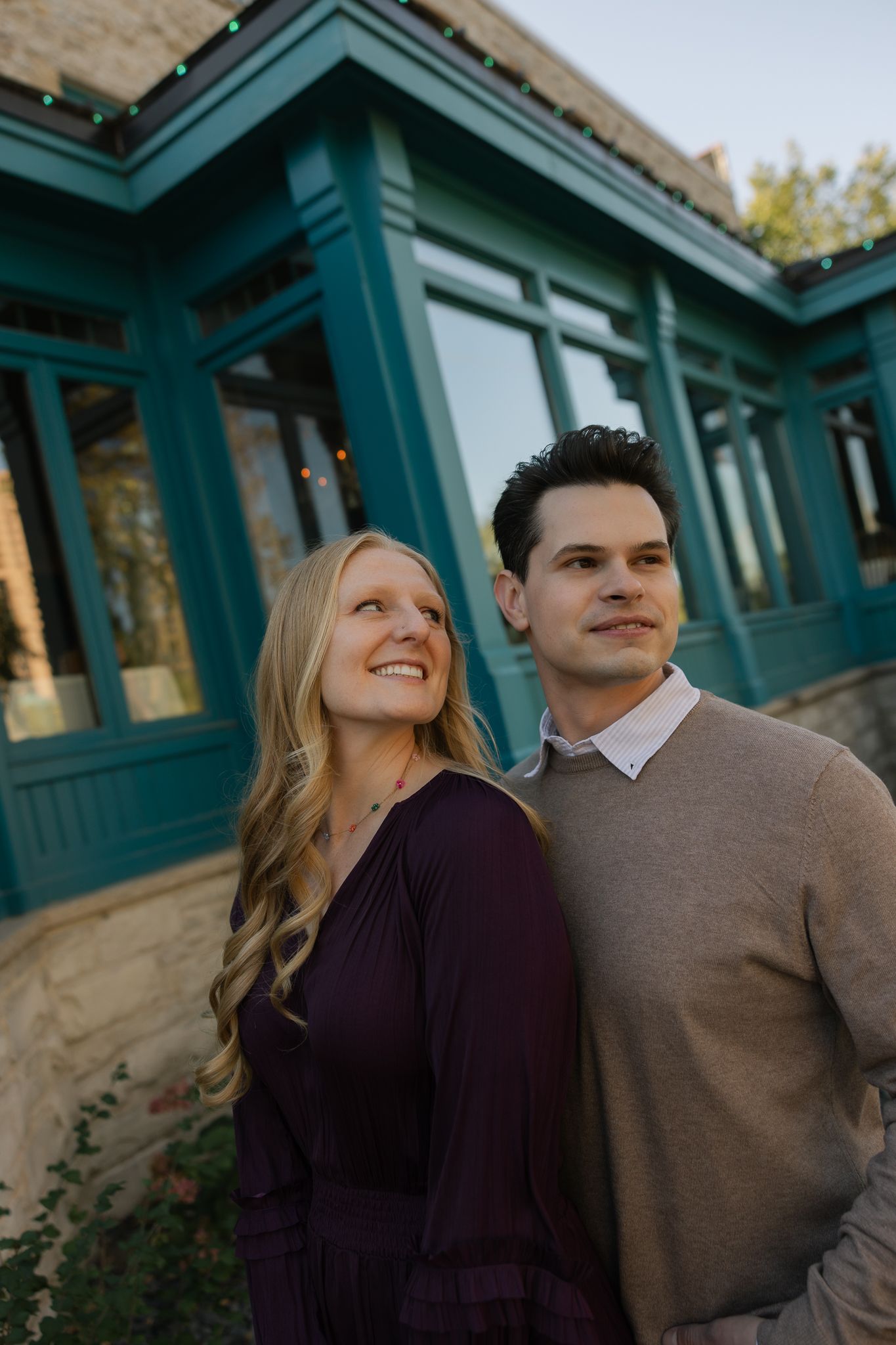 Couple in purple and tan sweaters sharing loving glances against teal-trimmed building.