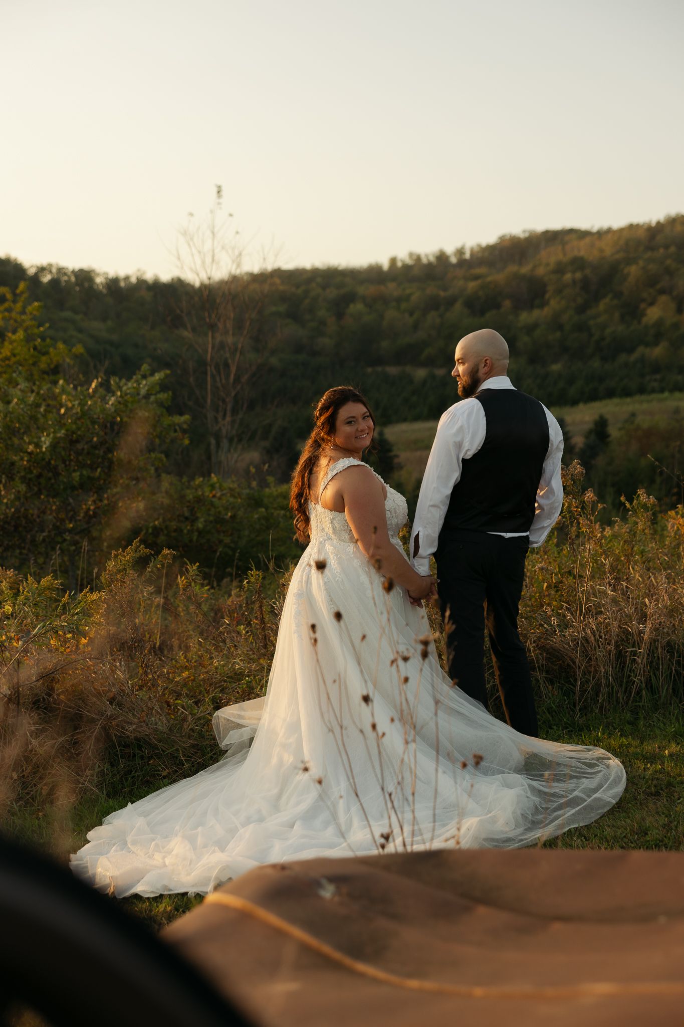 Romantic sunset scene with couple in wedding attire against rolling hills backdrop.