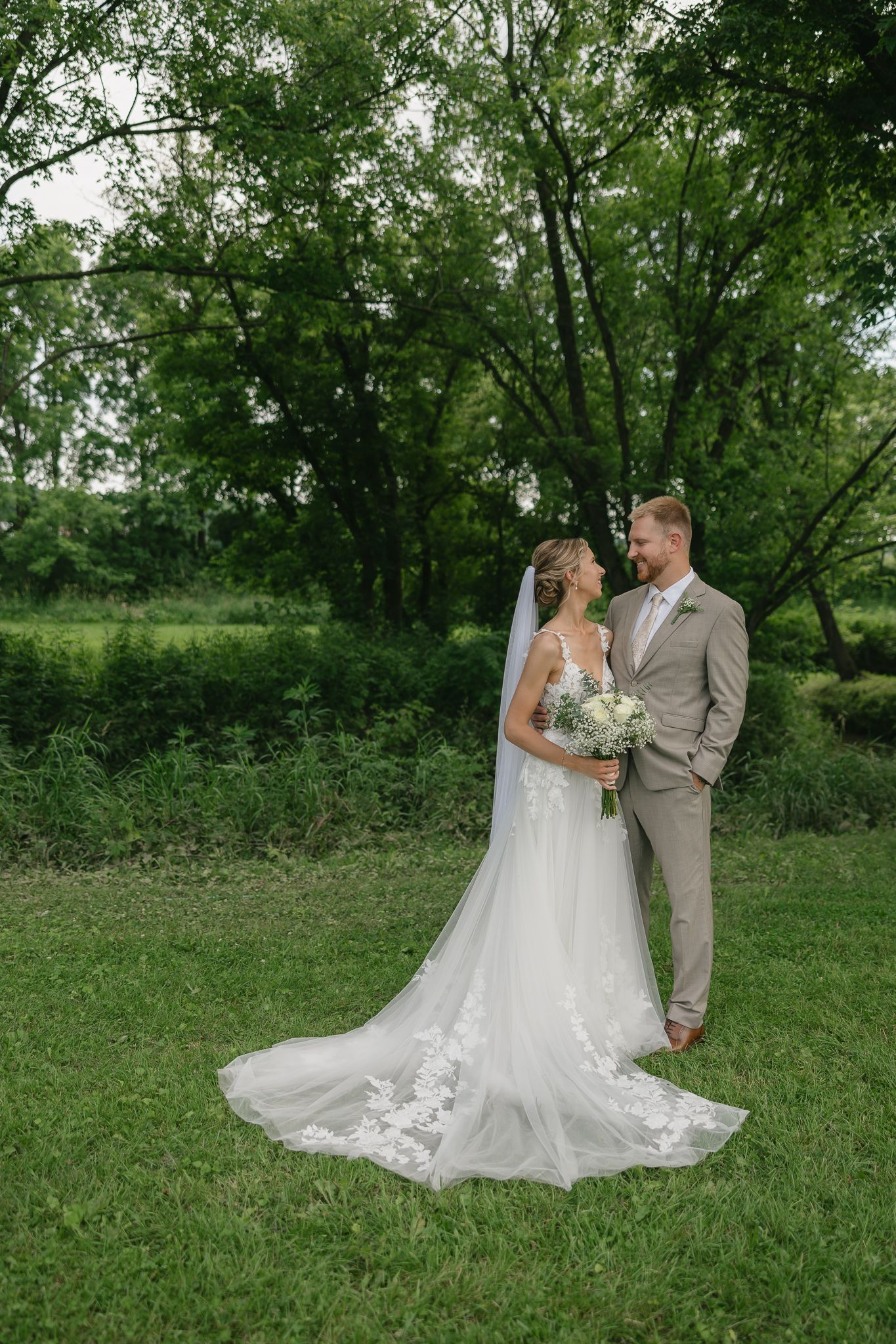 Wedding couple poses for portraits in a lush green outdoor setting with flowing white dress.