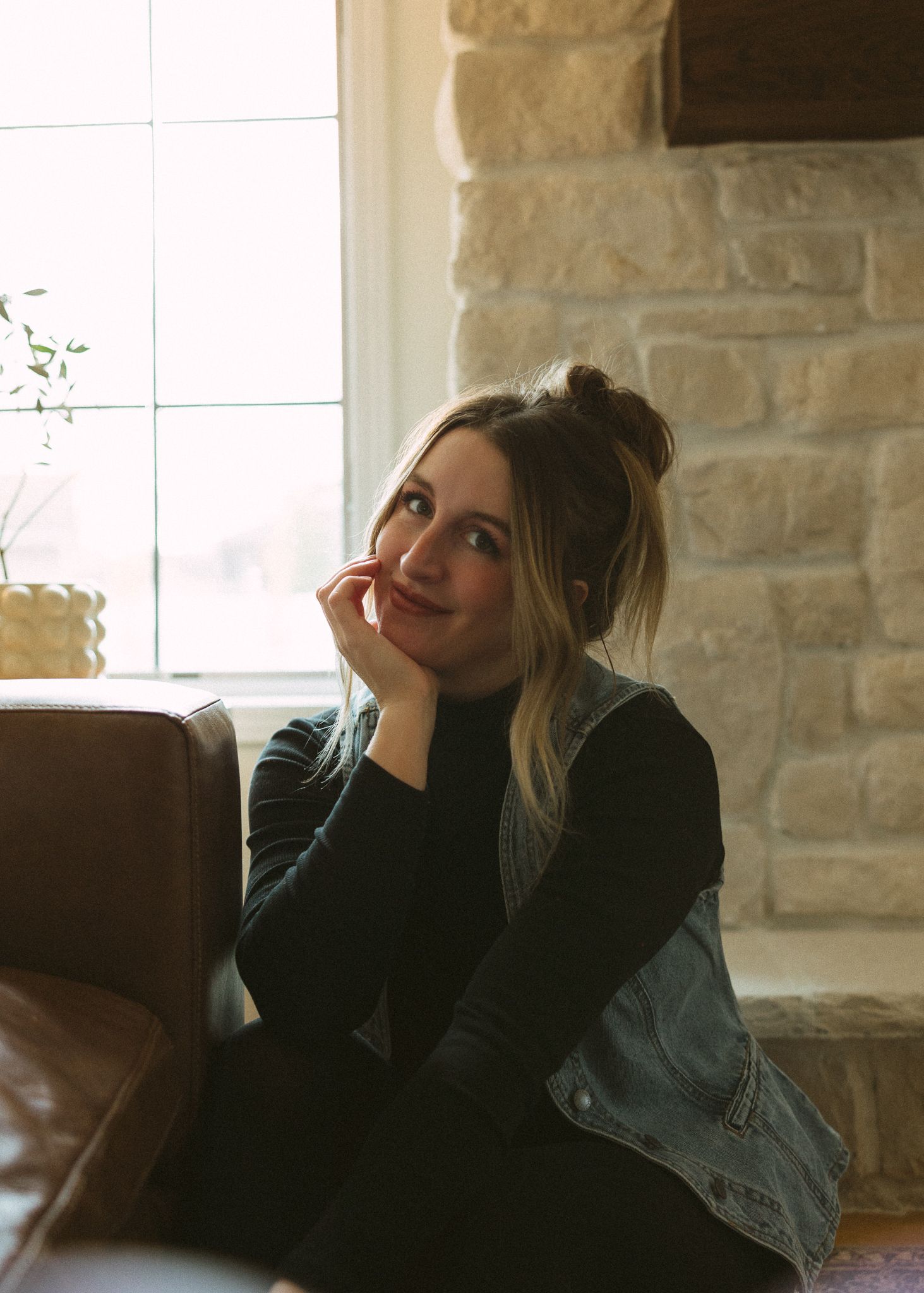 Thoughtful figure in denim jacket and black sweater rests chin on hand while sitting on leather couch near stone wall.