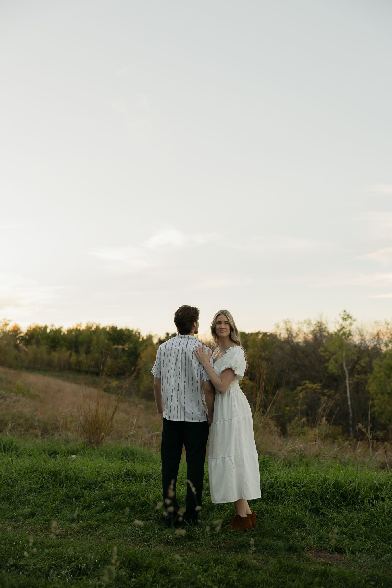 Couple in white and striped outfits standing together at sunset on grassy hillside.
