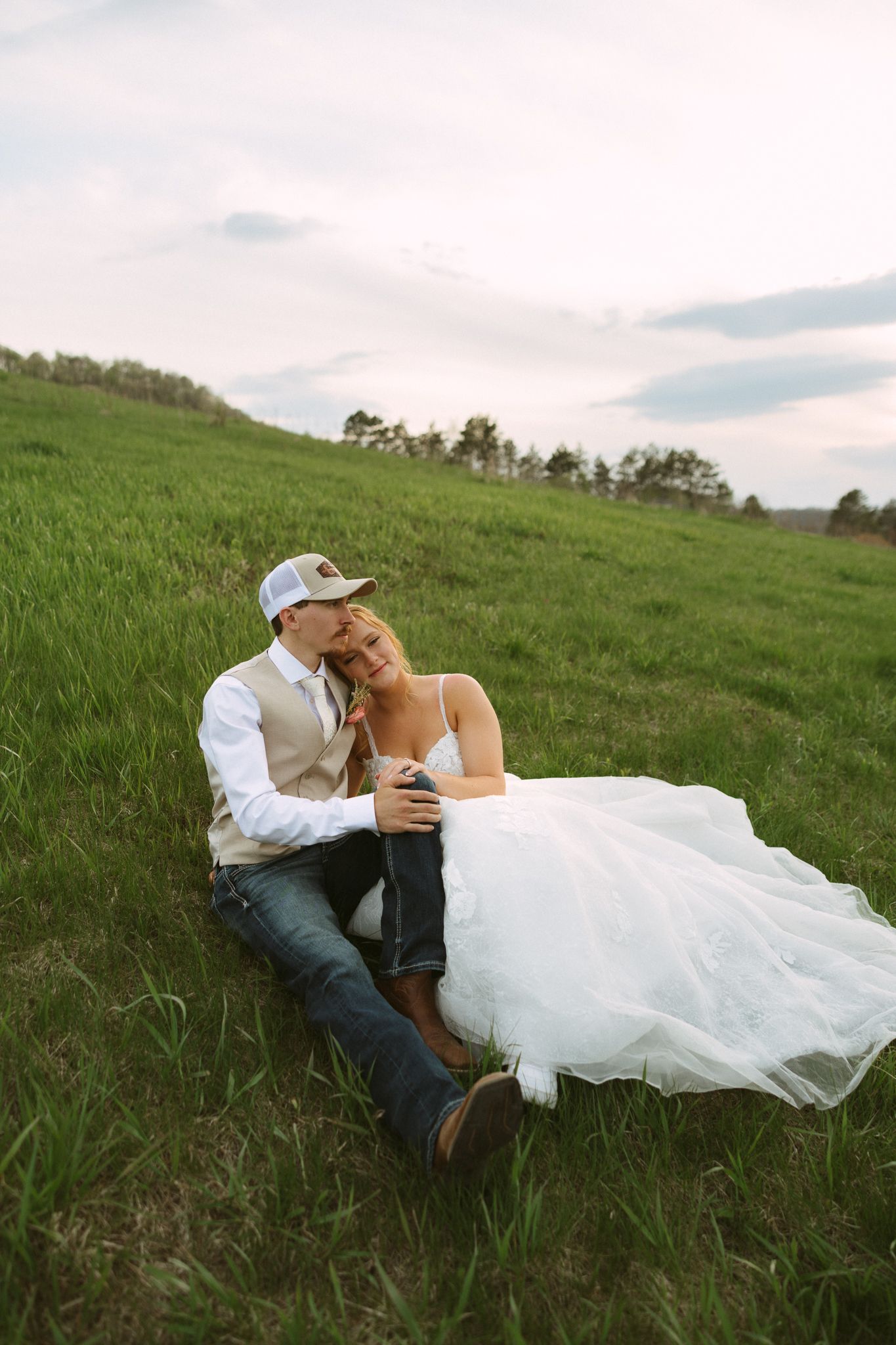 Bride and groom sitting on a green hillside, bride in white gown, groom in vest and cap, sharing a romantic moment.