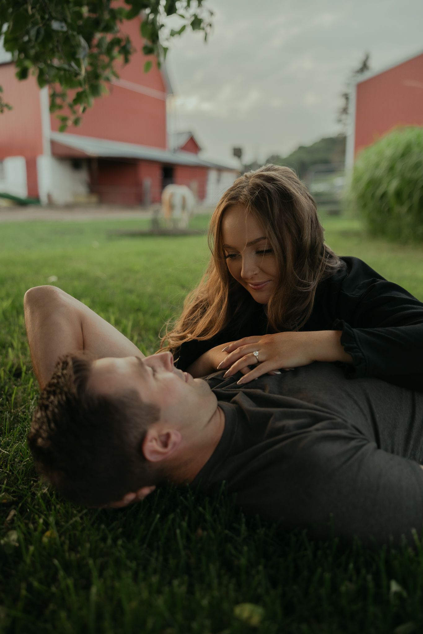 A couple shares a tender moment on grass with red barns in the background.