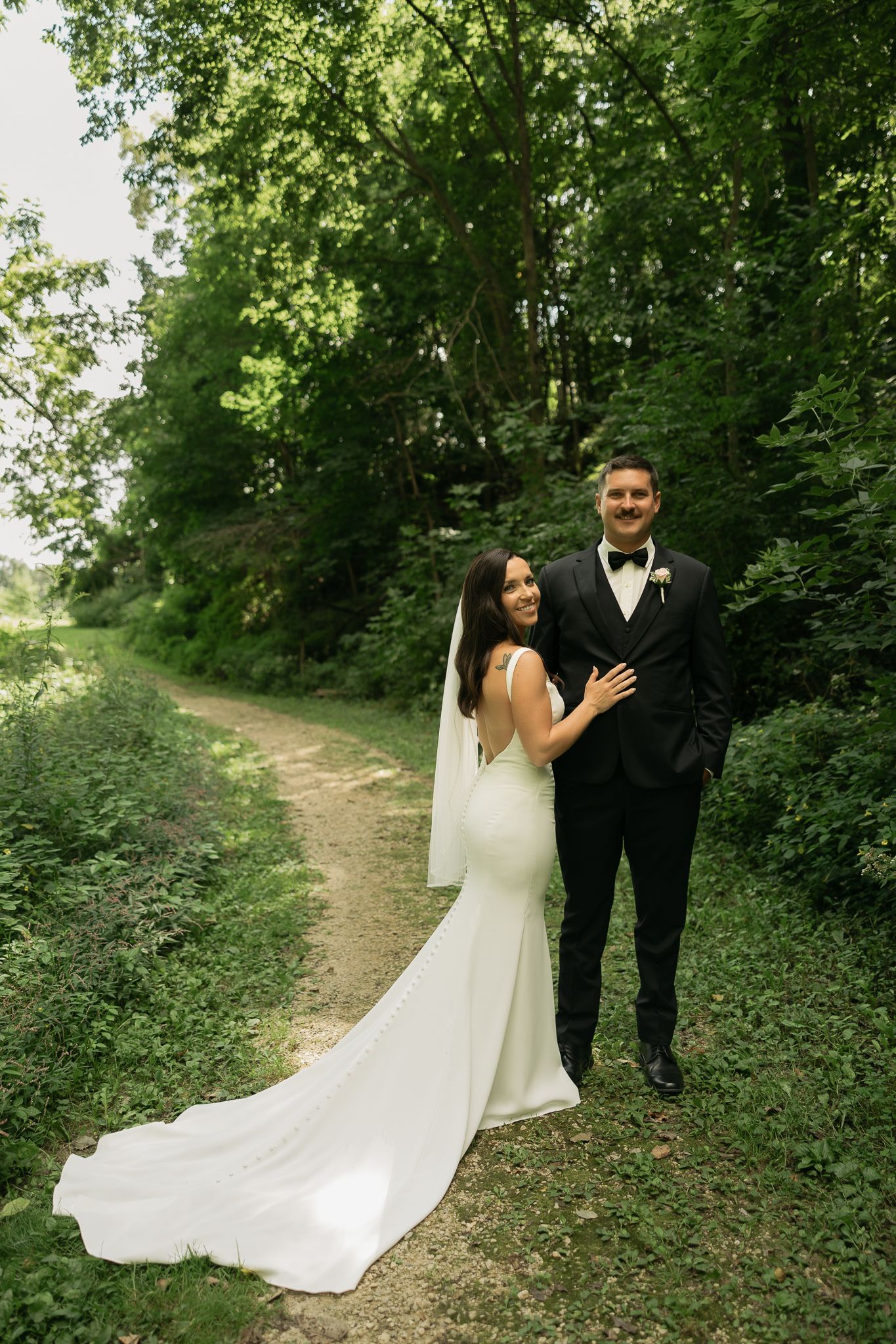 Bride in fitted white gown and groom pose on forest path with trailing veil.