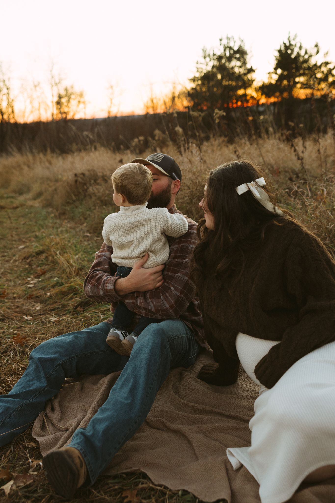 A person and two dogs sit together watching a golden sunset in a field of tall grass.