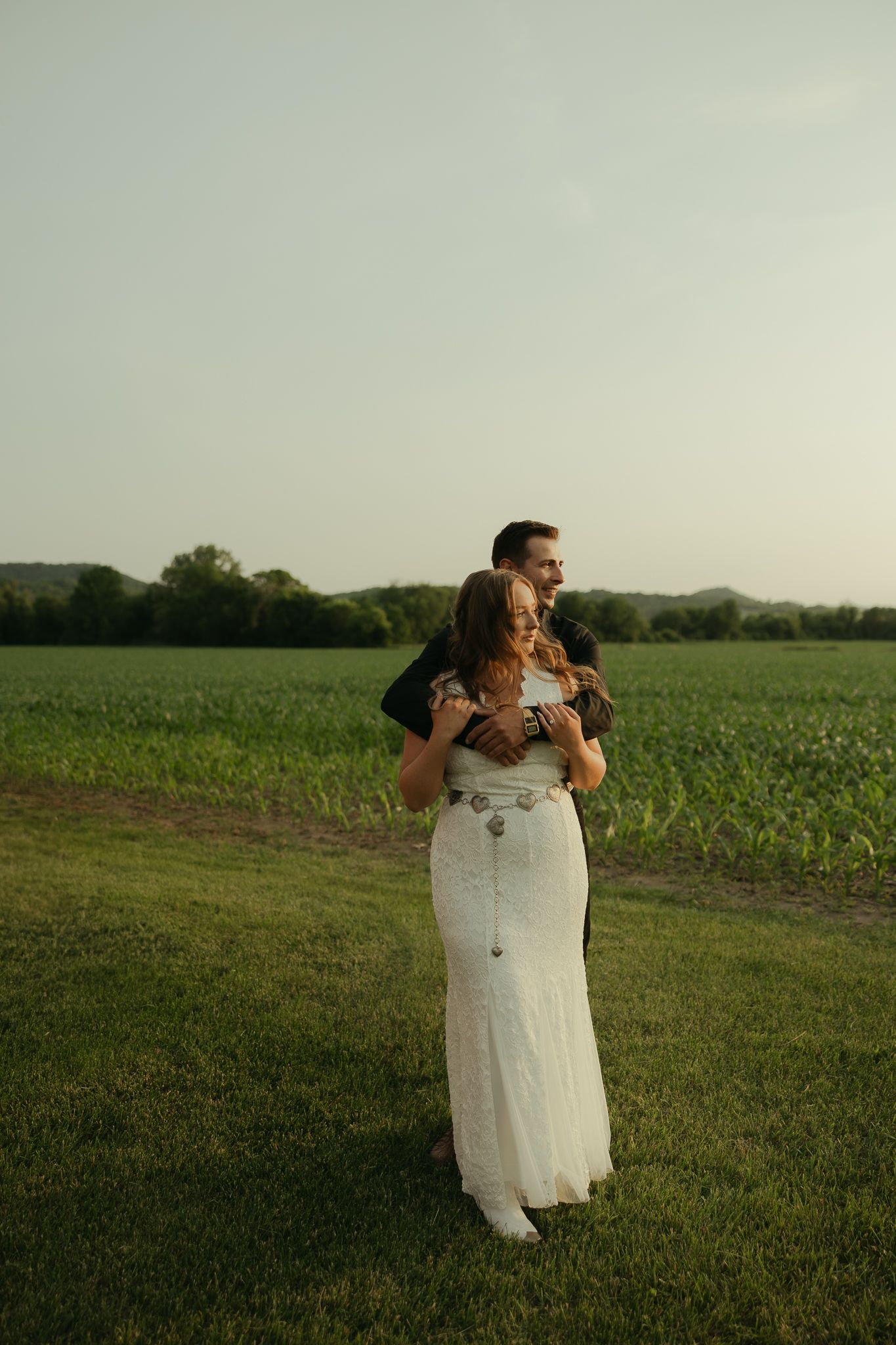 A couple in wedding attire shares an intimate moment in a green field at sunset.
