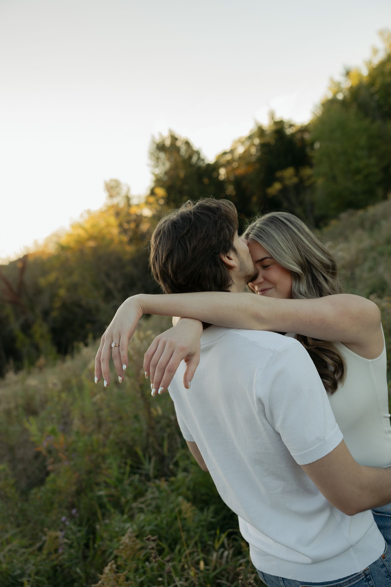 A romantic embrace between a couple in a white shirt and casual attire against a sunset-lit natural backdrop.