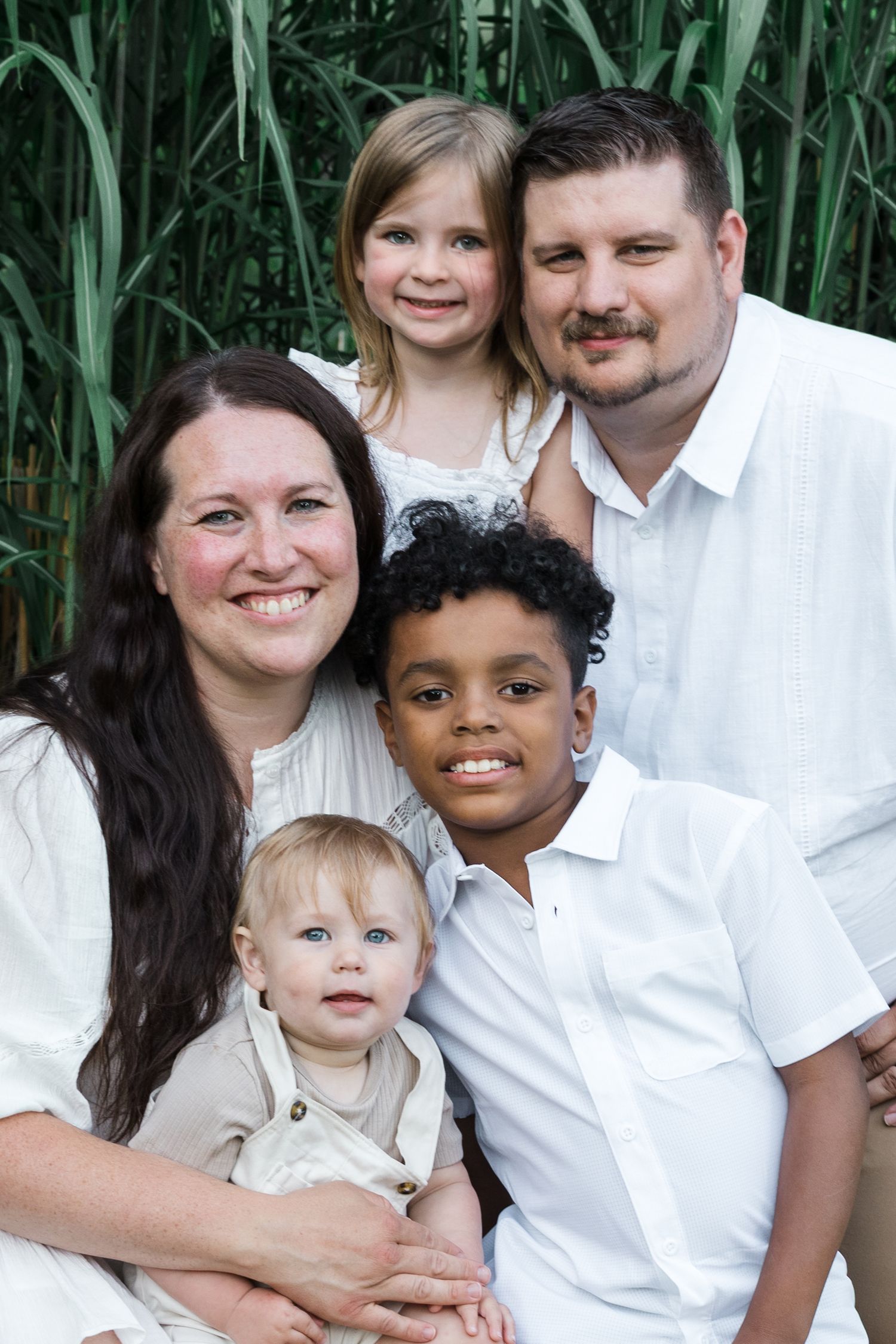 Happy family of five posing outdoors in white outfits, with green foliage in the background.