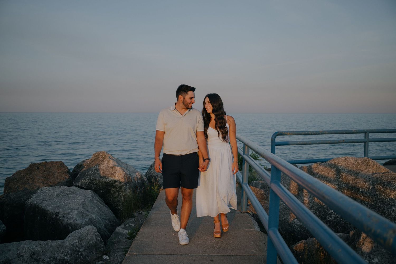 A couple walks along a coastal path during sunset holding hands with ocean views in the background on Lake Huron engagement session