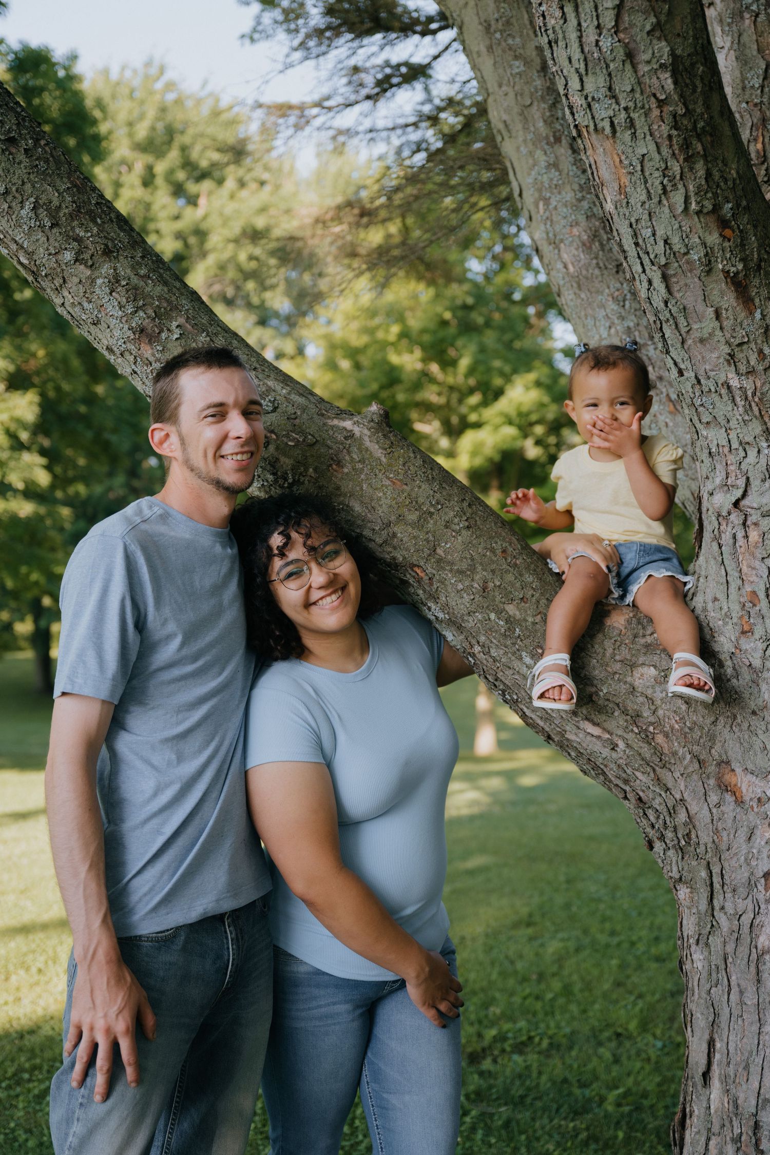 A couple shares playful and affectionate moments while posing near a large tree trunk in a sunny park setting.