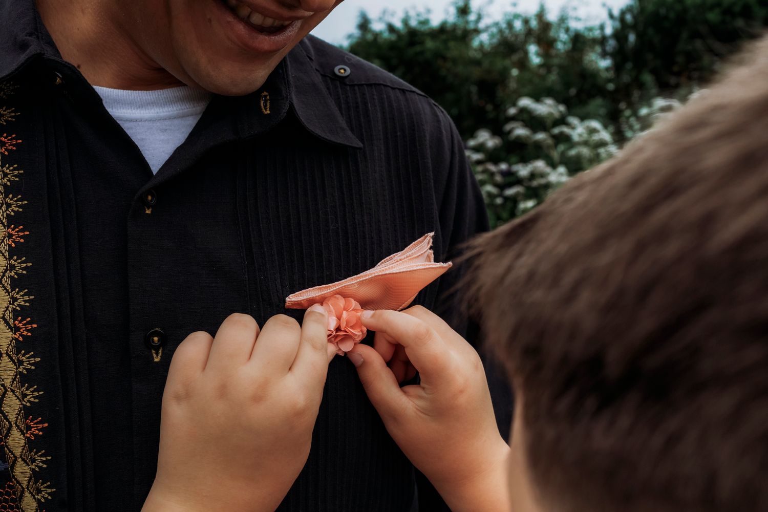 Child pinning a pink boutonniere flower onto a smiling man in a black shirt outdoors.