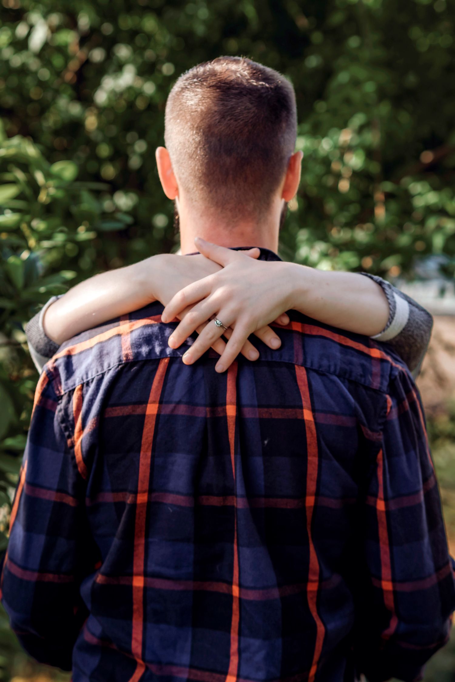 A person in a plaid shirt seen from behind with someone's hand resting on their shoulder, wearing a ring.