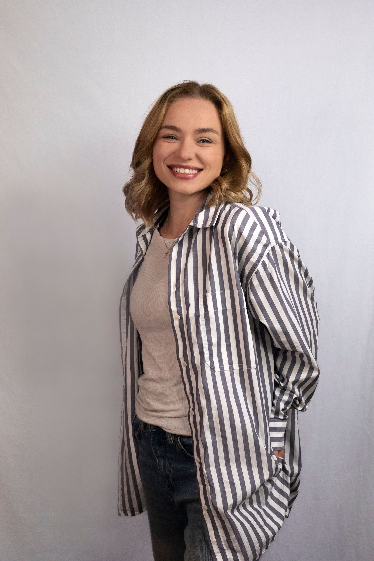 Smiling woman in striped oversized shirt layered over beige top and jeans, posing against white background.