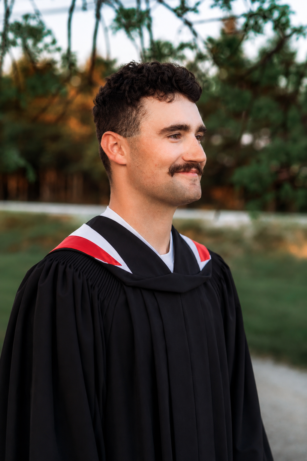 Young man in black graduation gown with red and white hood smiling outdoors at graduation ceremony.