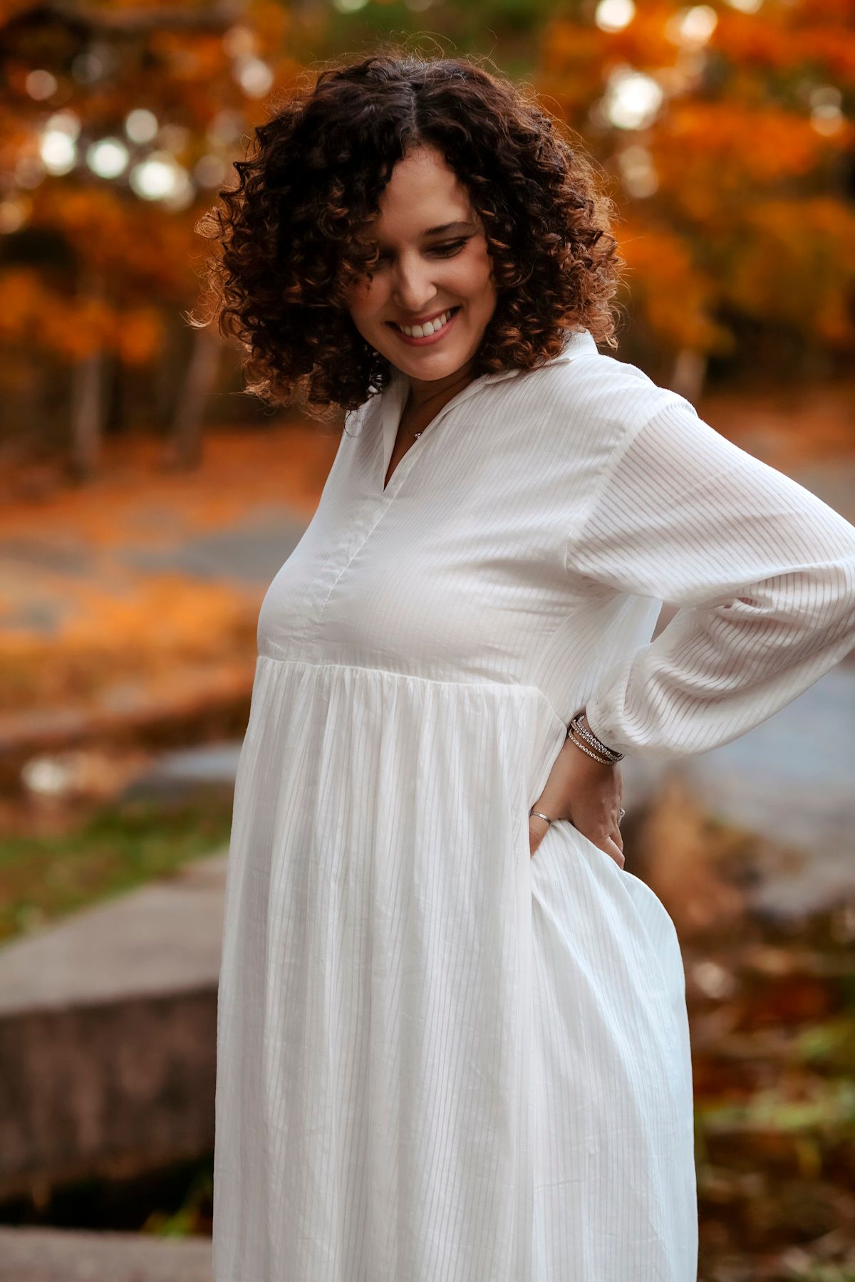 Woman in white dress smiling outdoors among autumn foliage.