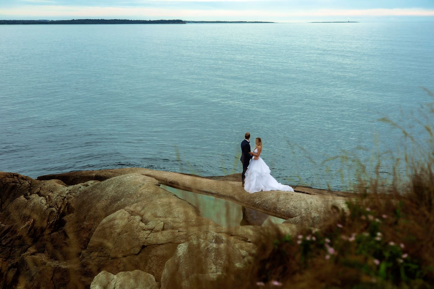 Bride and groom standing on coastal rocks overlooking calm blue ocean during romantic wedding photo session.