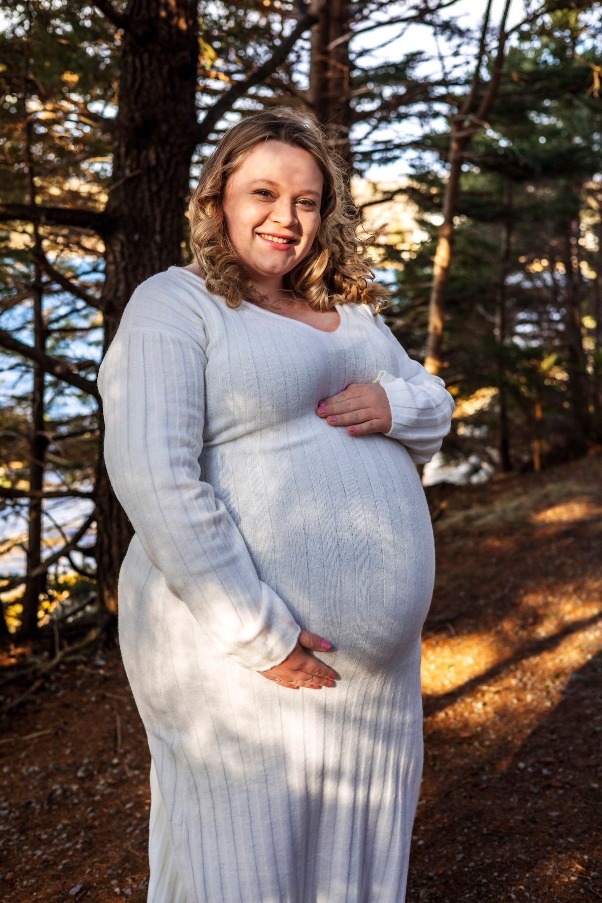 Smiling pregnant woman in white ribbed dress cradling her baby bump outdoors among pine trees in warm sunlight.