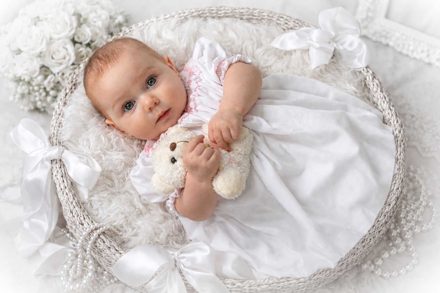 Baby in white dress holding teddy bear in wicker basket with white bows and floral decorations Halifax newborn photographer