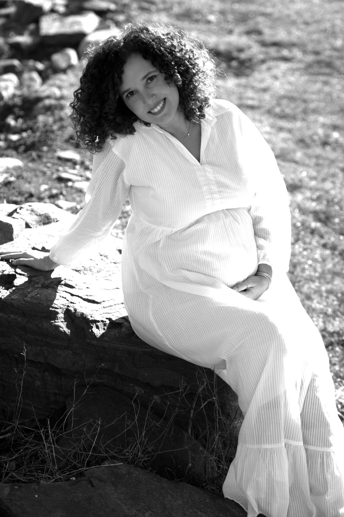 Smiling pregnant woman in white dress sitting on rocks outdoors, black and white maternity portrait photo.