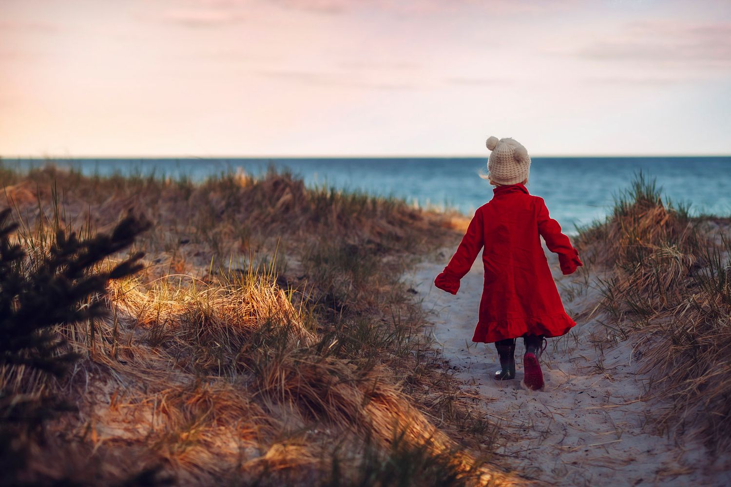 Young child in red coat and white hat walking toward ocean through sandy dunes at sunset.