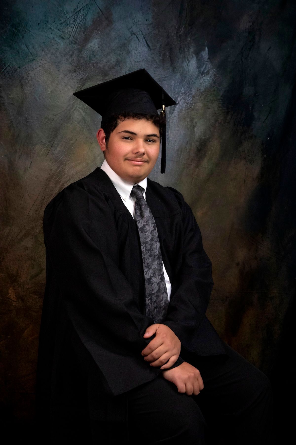 Young man in black graduation gown and mortarboard cap smiling for formal portrait photo.