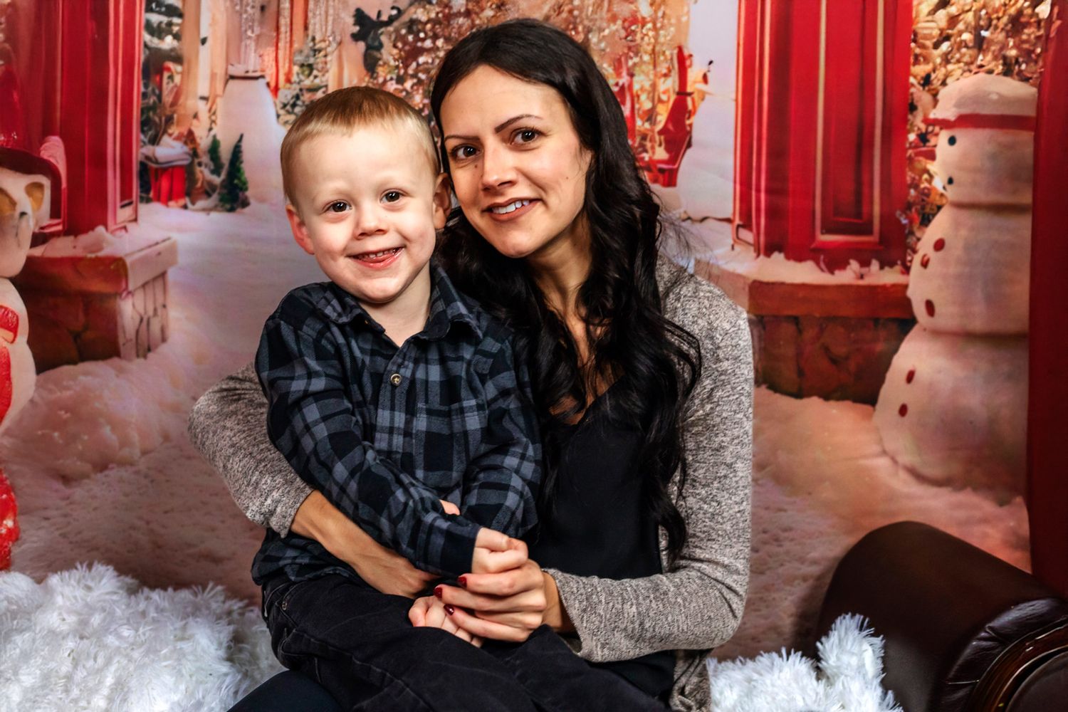Smiling woman with long dark hair hugs a young blond boy in a plaid shirt in front of a festive Christmas backdrop with a snowman.