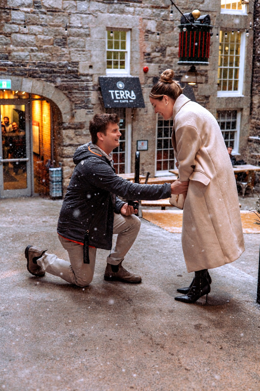 A man kneels on one knee proposing to a woman in a beige coat as snow falls outside a stone cafe in a historic courtyard.