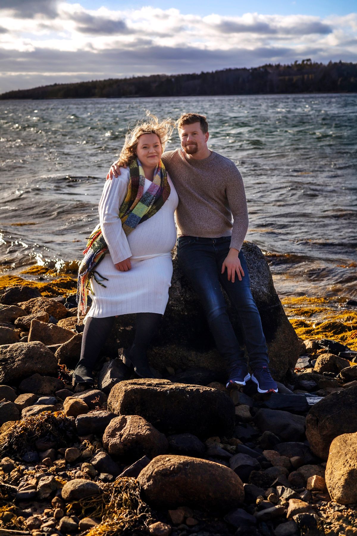 Pregnant couple sitting on rocky shoreline with choppy water and cloudy sky behind them.
