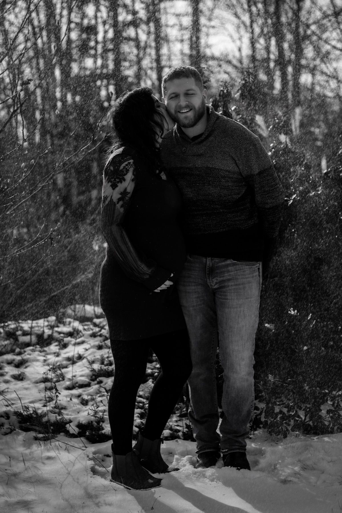 Black and white photo of a smiling couple embracing in a snowy winter forest, woman leaning into mans shoulder.