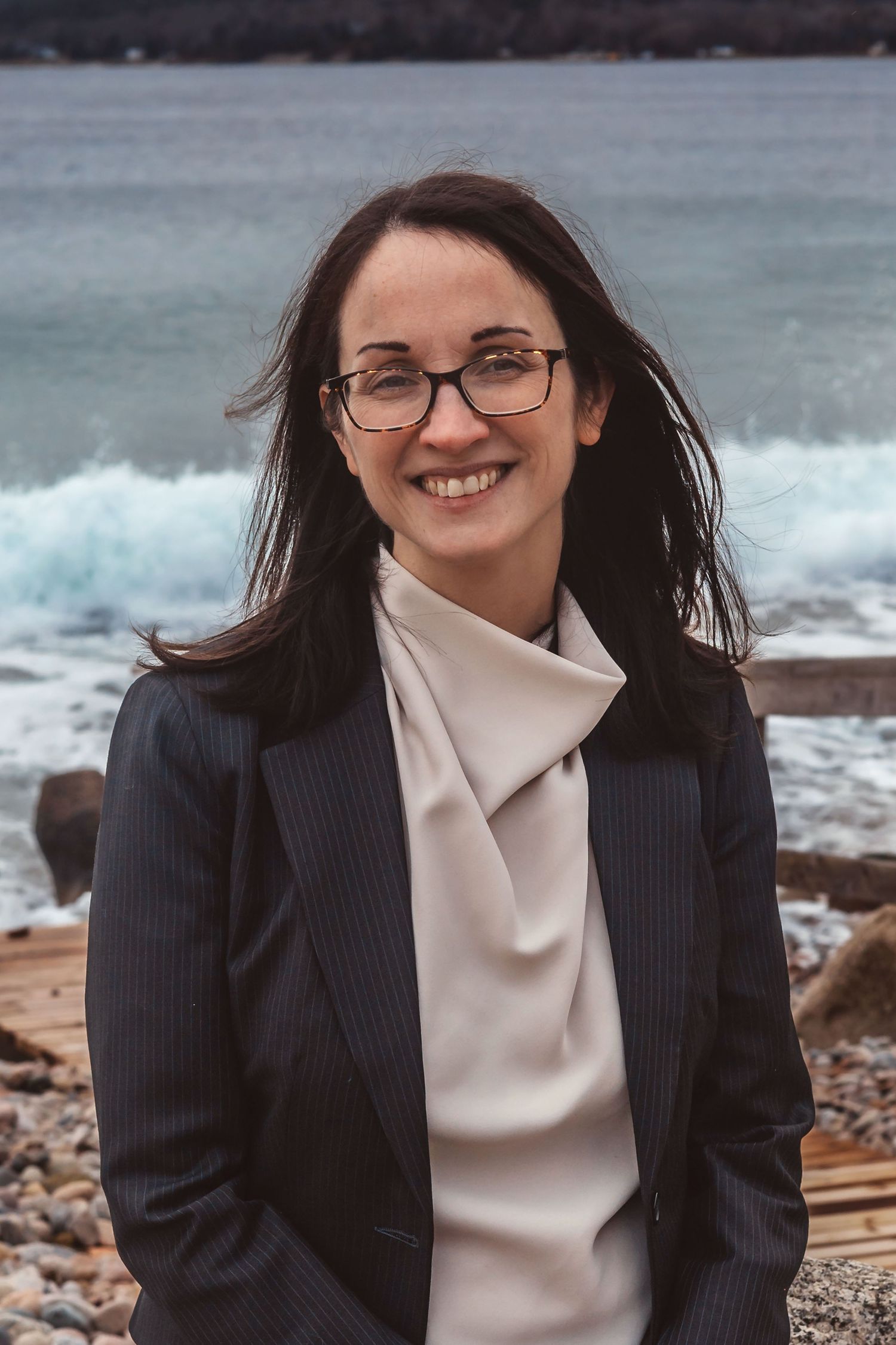 Smiling woman with glasses and dark hair in a blazer poses on a rocky beach with ocean waves behind her.