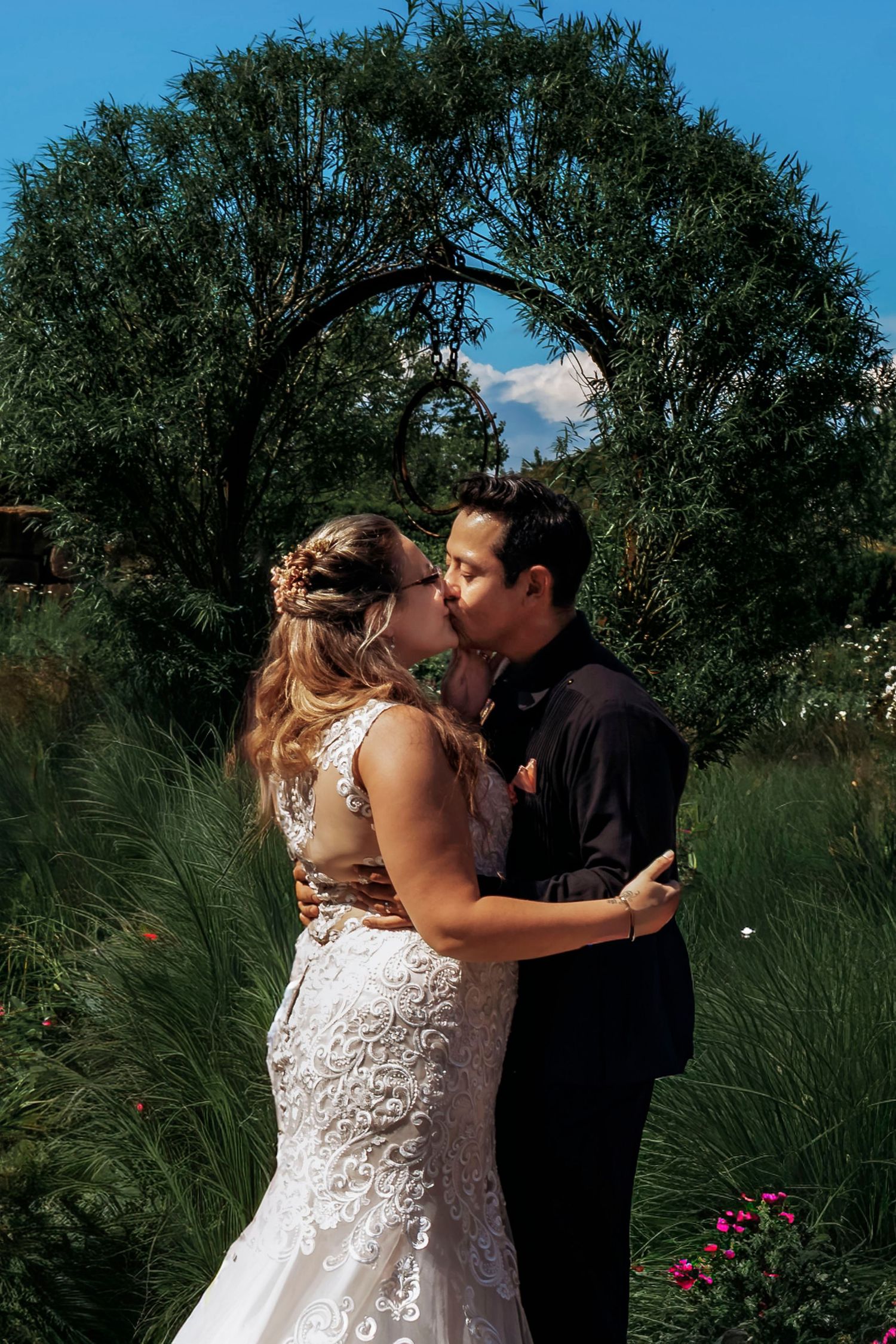 A bride and groom share their first kiss at an outdoor wedding ceremony surrounded by lush green gardens under a blue sky.