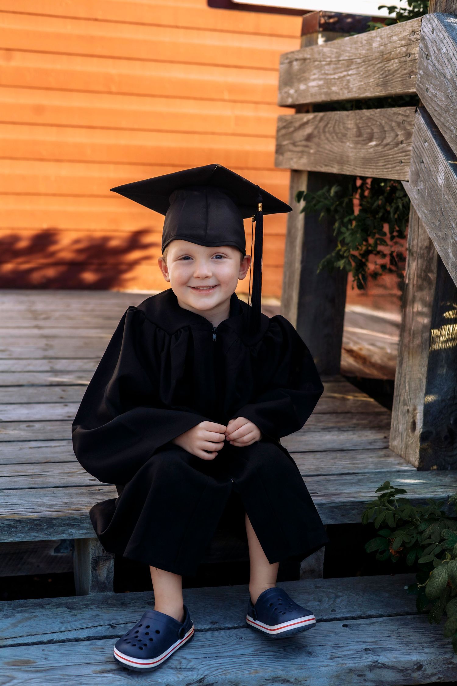 Young child smiling in black graduation cap and gown, sitting on wooden steps in blue Crocs shoes.