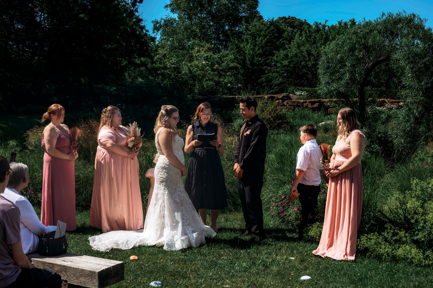 Outdoor wedding ceremony with bride, groom, bridesmaids in pink dresses, and officiant gathered on green lawn under sunny sky.