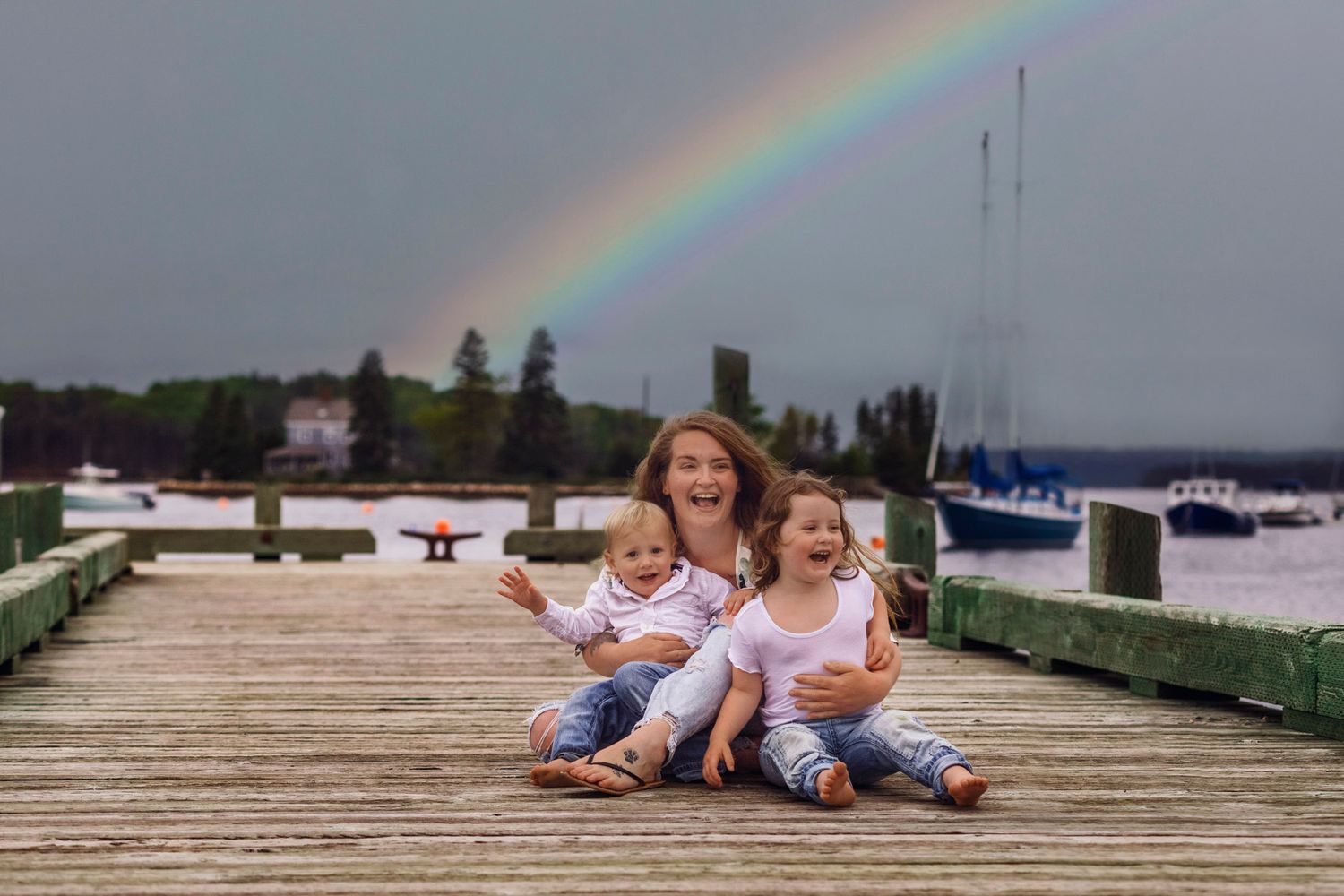 Three children sit smiling on a wooden dock with a vibrant rainbow arching over a calm harbor behind them.