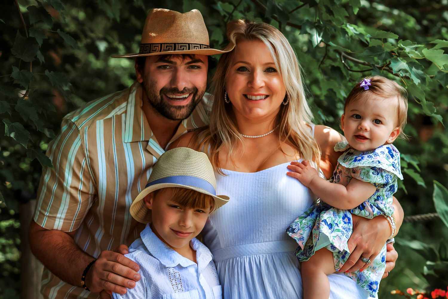 Happy family of four posing outdoors, parents with two young children wearing hats, surrounded by lush green foliage.