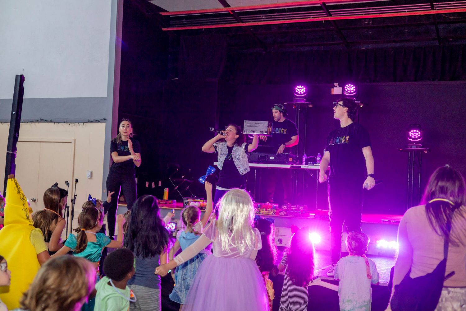 Children enjoy a live kids entertainment show on a colorful stage with performers singing and dancing under purple lights.