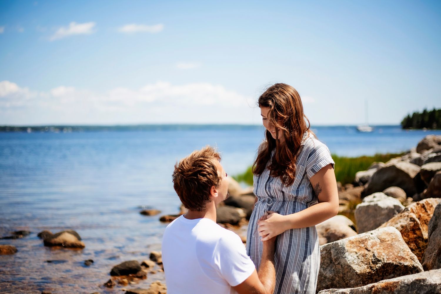 A couple shares a romantic moment by rocky shoreline on a sunny day, with calm blue water in the background.