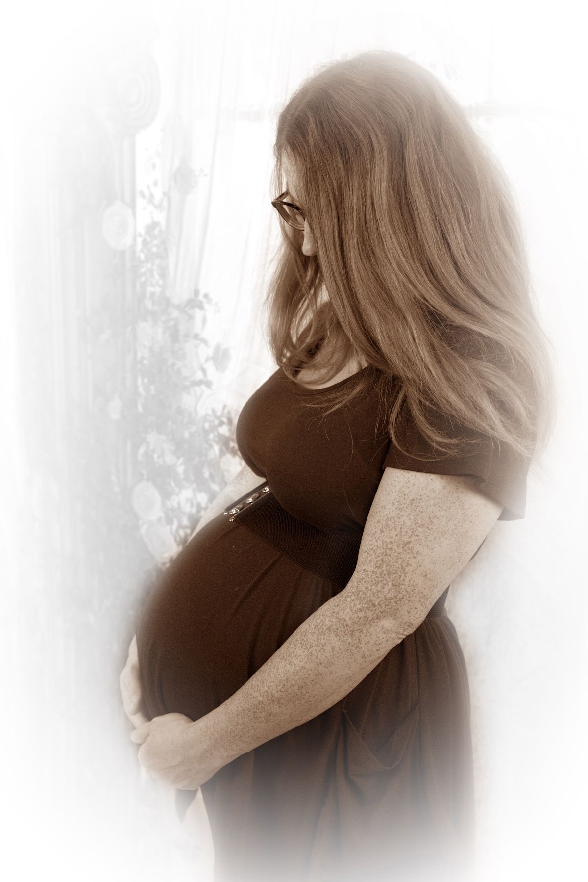 Pregnant woman in brown dress cradling her baby bump, sepia-toned maternity portrait near a window.