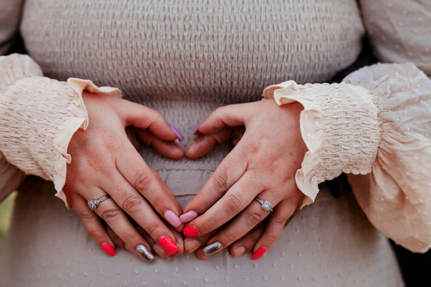 Pregnant woman forming a heart shape with her hands on her baby bump, wearing red nail polish and a ring.
