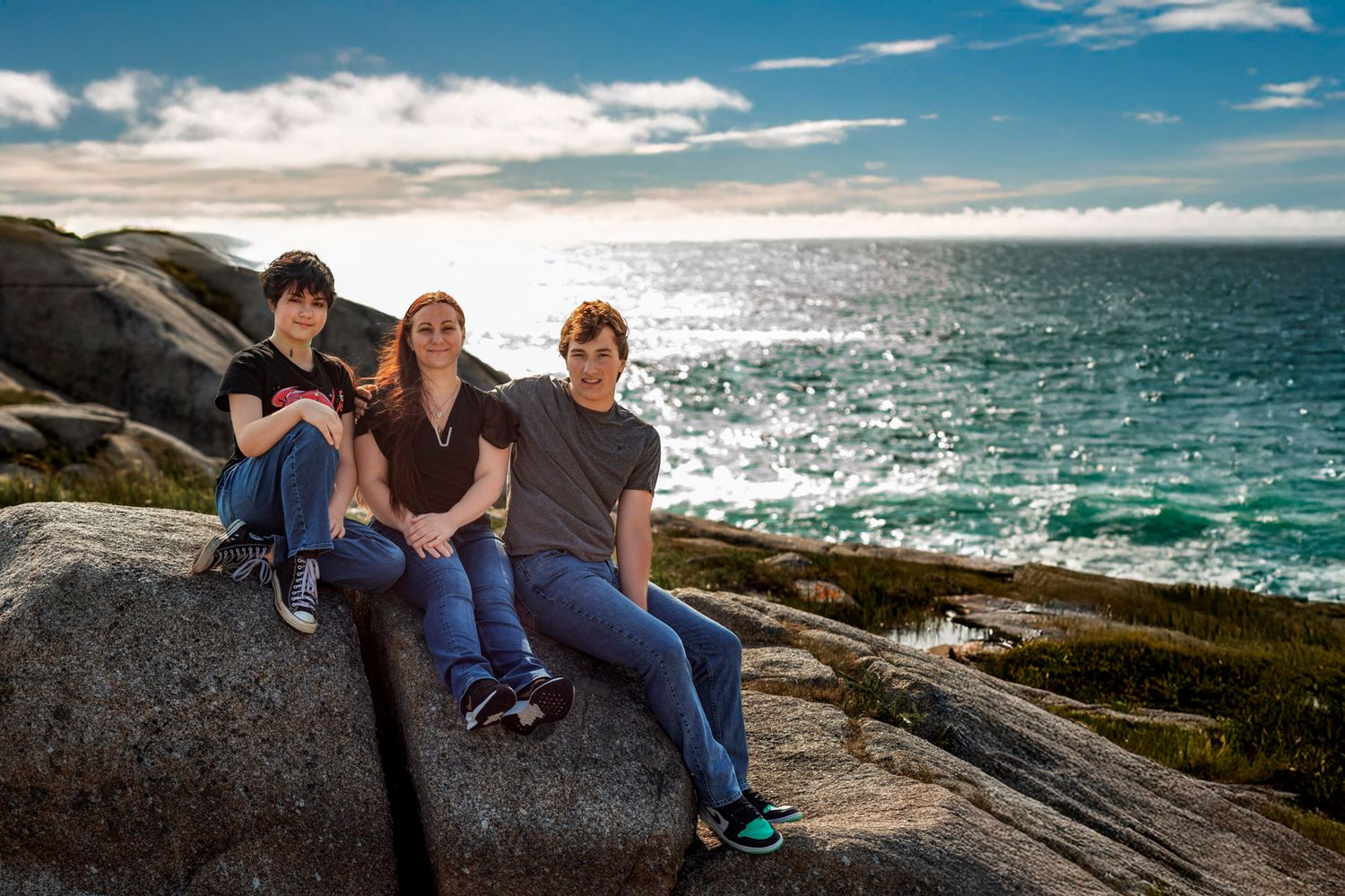 Three people sitting on coastal rocks with turquoise ocean and blue sky backdrop in a scenic outdoor portrait.
