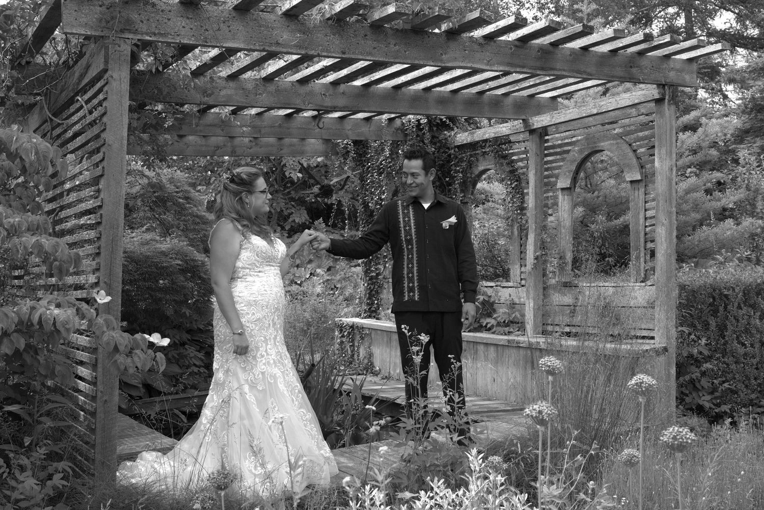 Black and white photo of a bride and groom holding hands under a wooden pergola in a lush garden wedding setting.