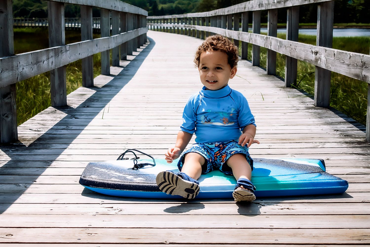 Smiling toddler in blue rashguard sits on a surfboard on a wooden boardwalk near a lake on a sunny day.