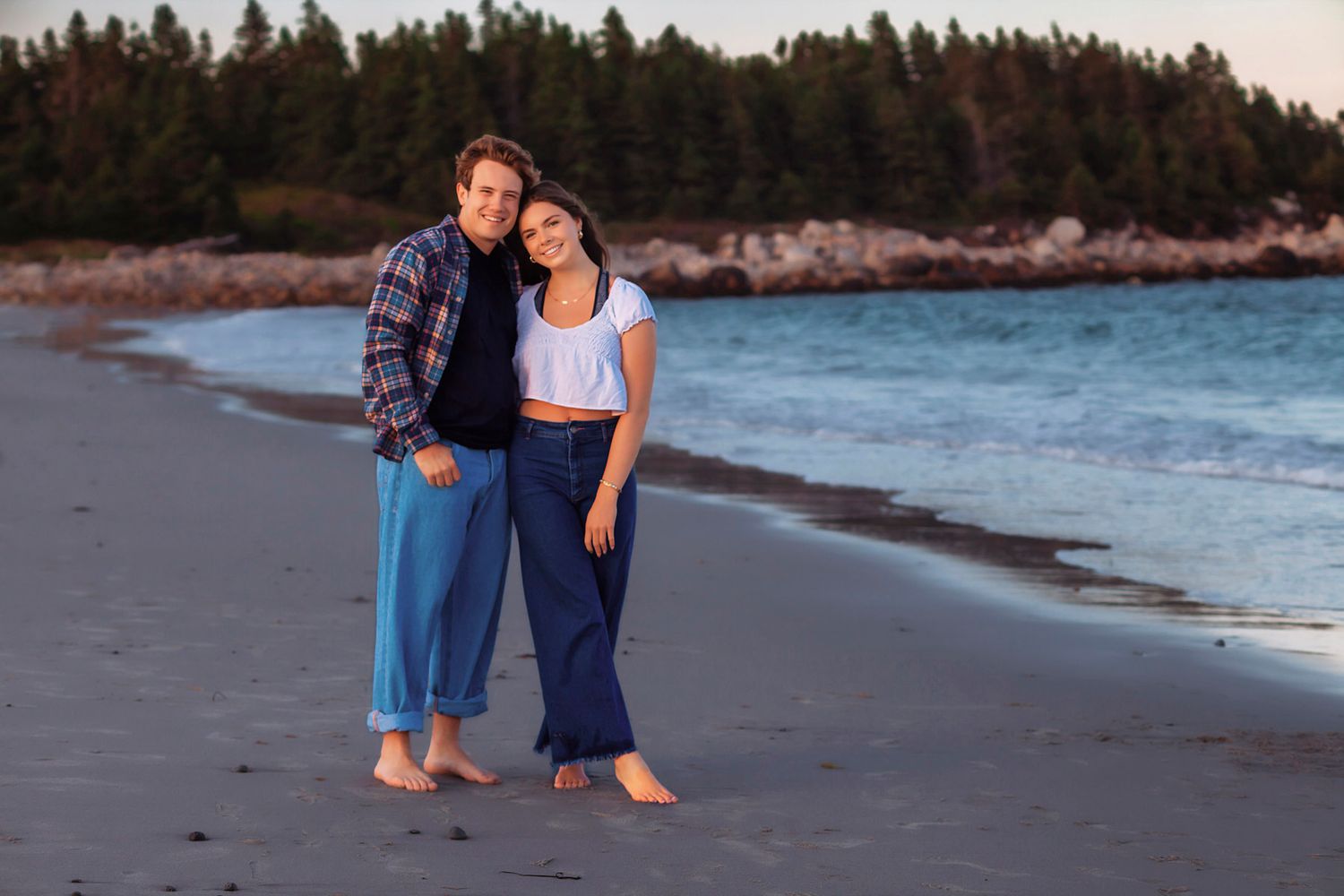 Couple standing barefoot on a sandy beach at sunset, wearing blue pants, with turquoise water and pine trees behind them.