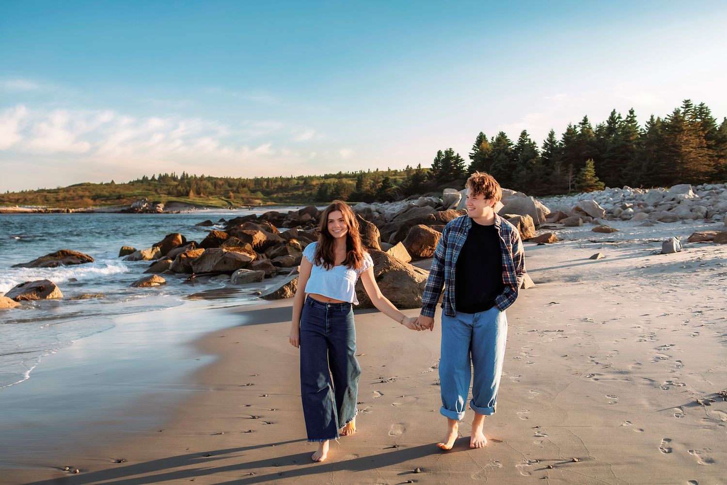 newly engagement shoot in Halifax couple walk hand-in-hand barefoot on a sandy beach at sunset with rocky shores and pine trees behind them.