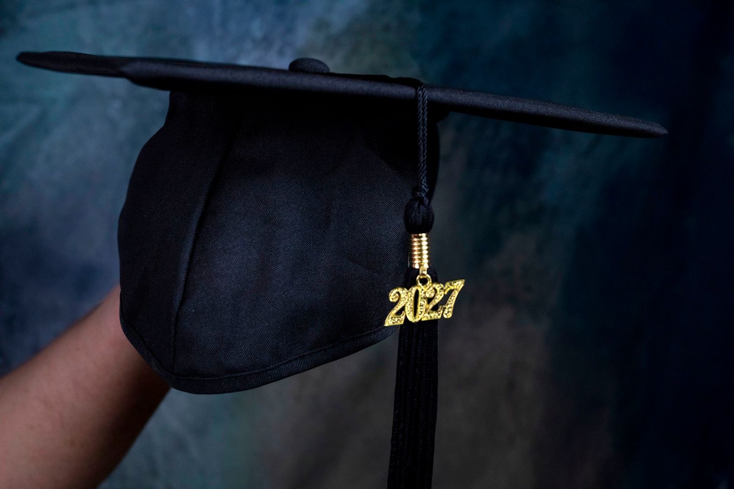 Hand holding a black 2027 graduation cap with gold tassel charm against a dark background during a studio session at Atlantic Memories