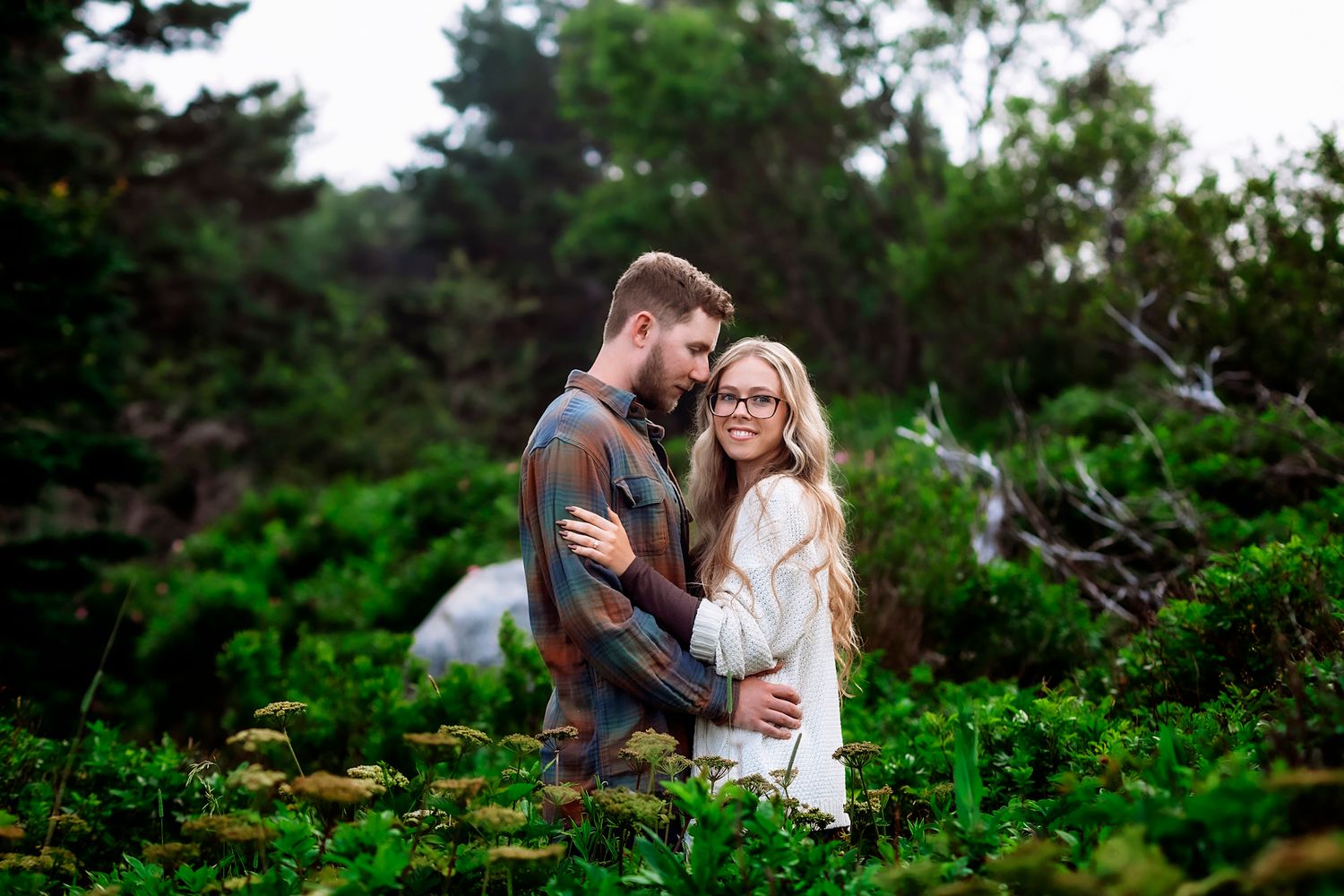 Couple embracing in lush green garden, woman with glasses and long blonde hair, man in plaid shirt.