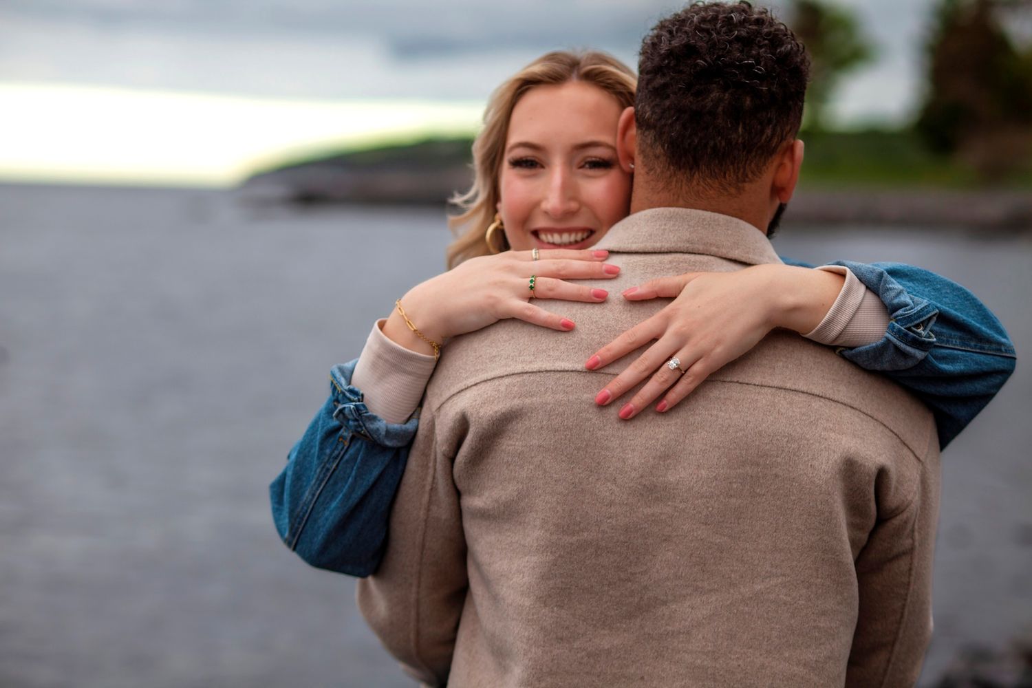 Happy blonde woman hugging a man near a lake at dusk, smiling warmly in a romantic outdoor embrace.