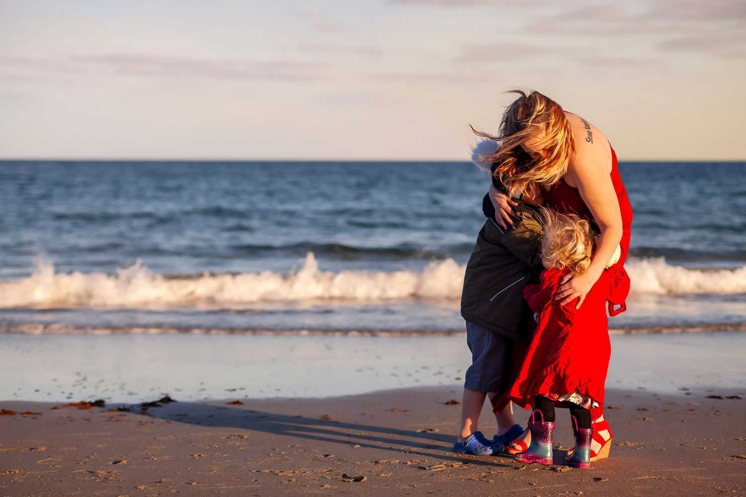 A mother hugging her two children on a sandy beach at sunset with ocean waves in the background.