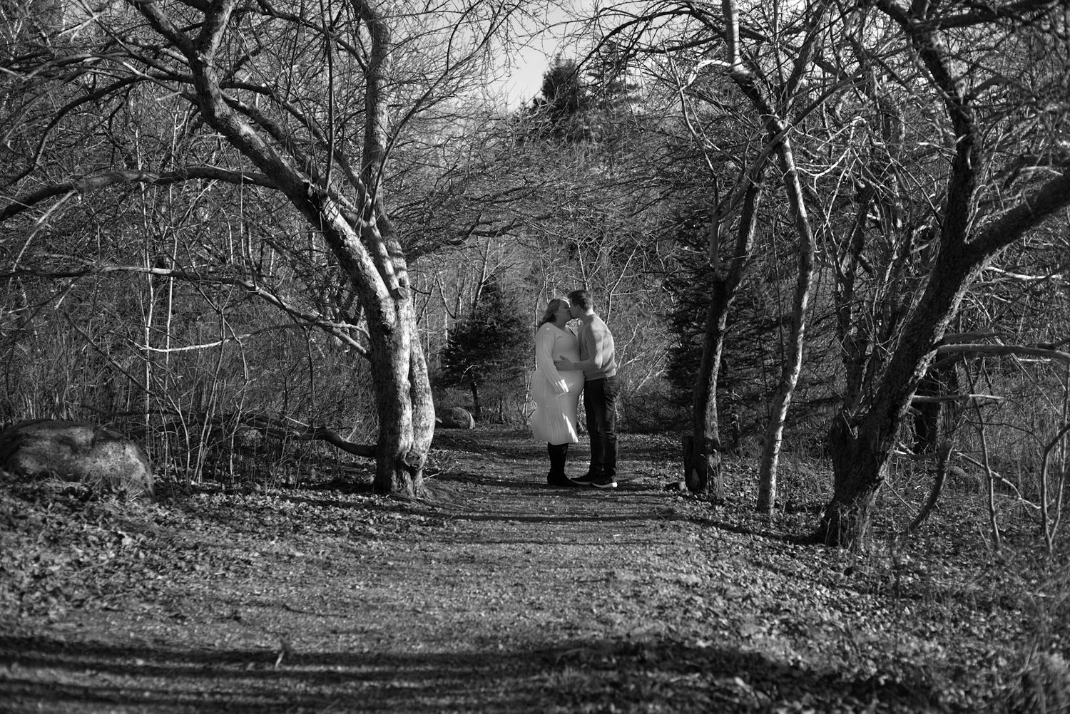 Black and white photo of a couple walking on a wooded path surrounded by bare trees in winter.